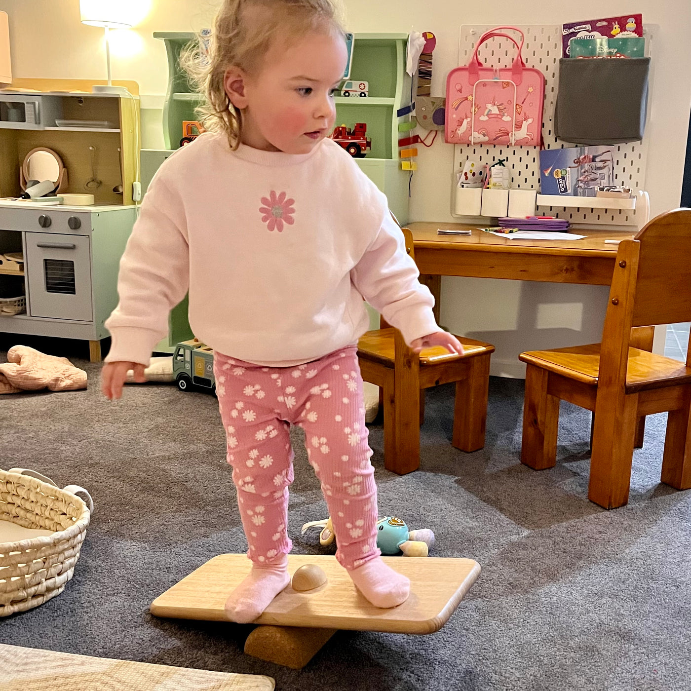 Child standing on a balance board in a playroom with toys and furniture.