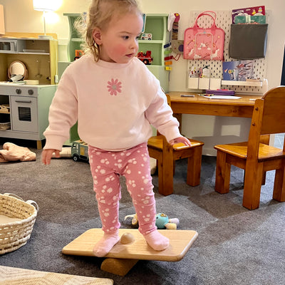 Child standing on a balance board in a playroom with toys and furniture.